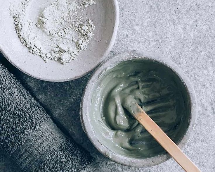 Two bowls containing different textured substances, possibly facial masks or scrubs, with an applicator in one bowl, on a textured surface.