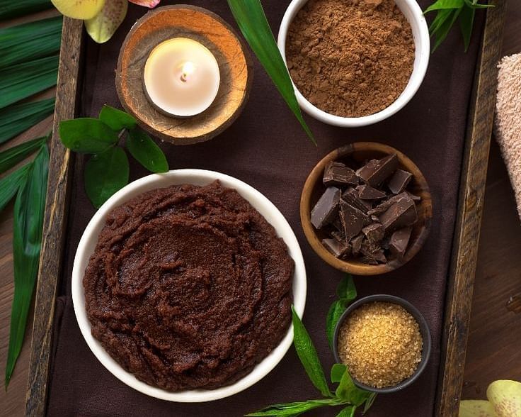 Flat lay of natural spa ingredients including cocoa, scrub powders, essential oils, and orchids arranged on a wooden tray.