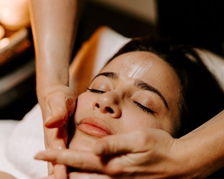 A person lying down receiving a facial treatment from two hands applying product to their face.