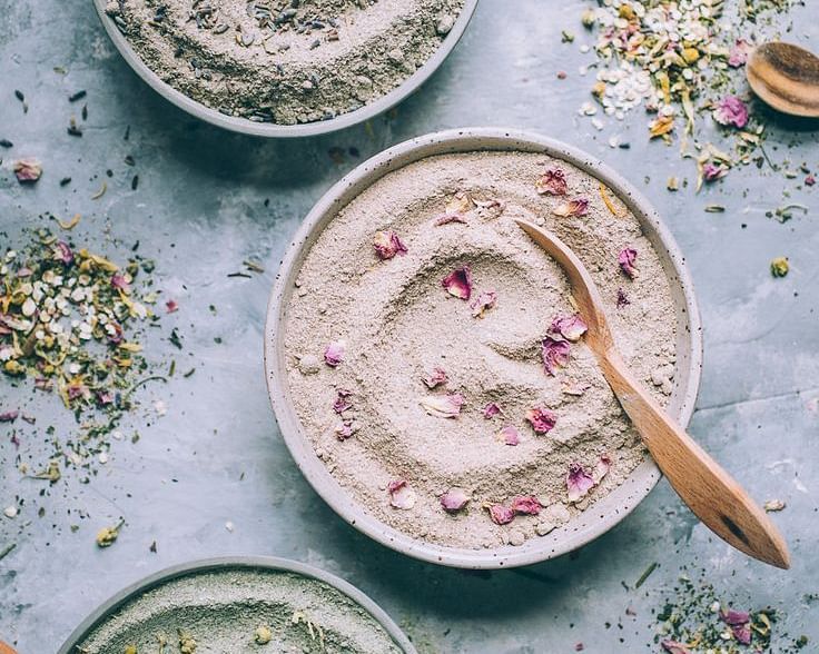 Flat lay of colorful face masks in bowls with wooden spoons, surrounded by herbs and flowers.