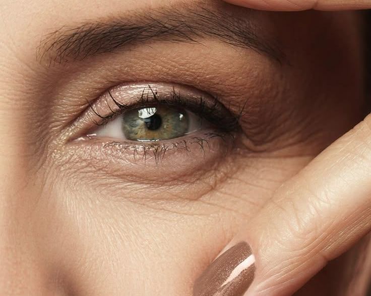A close-up of a person's eye and surrounding facial area, with fingers gently touching the skin.