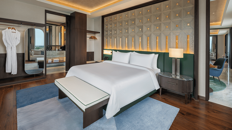 Executive Suite second bedroom with a double bed, padded bench, wooden floor, a bathrobe in an open wardrobe and a mirror at The Residency Signature Madurai.