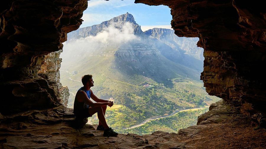 Photo of a man sitting inside a cave with hills in the background. 