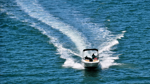 An aerial view depicts a motorboat speeding across open water, creating a wide wake, white foam, and rippling waves in its path.