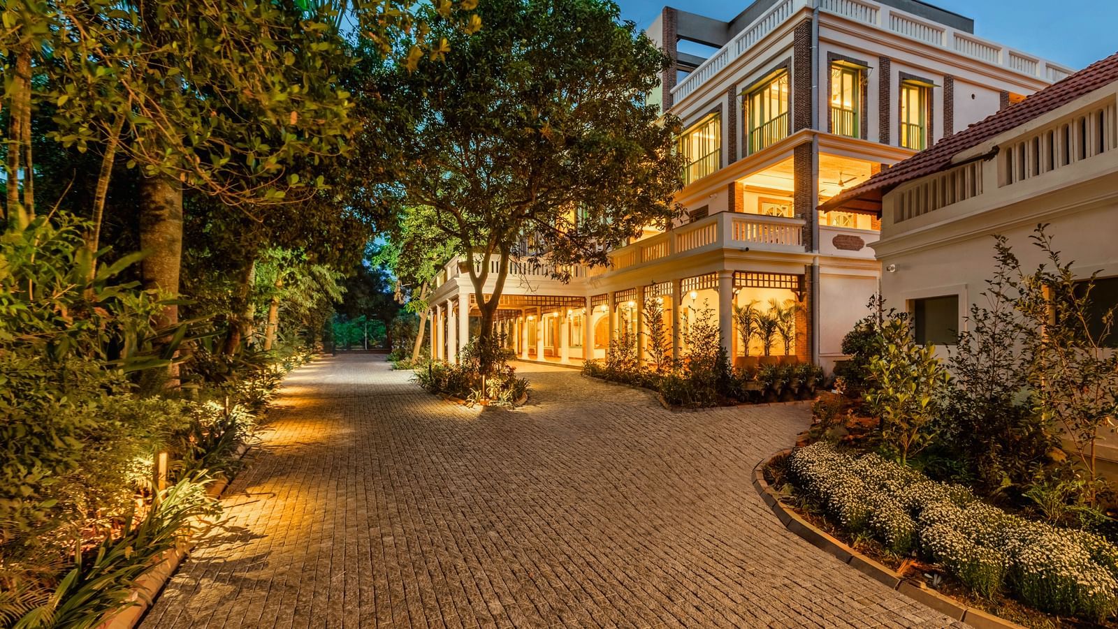 The illuminated colonial-style main building and lush, pebbled driveway at twilight at Mango Hill Auroville, Pondicherry.