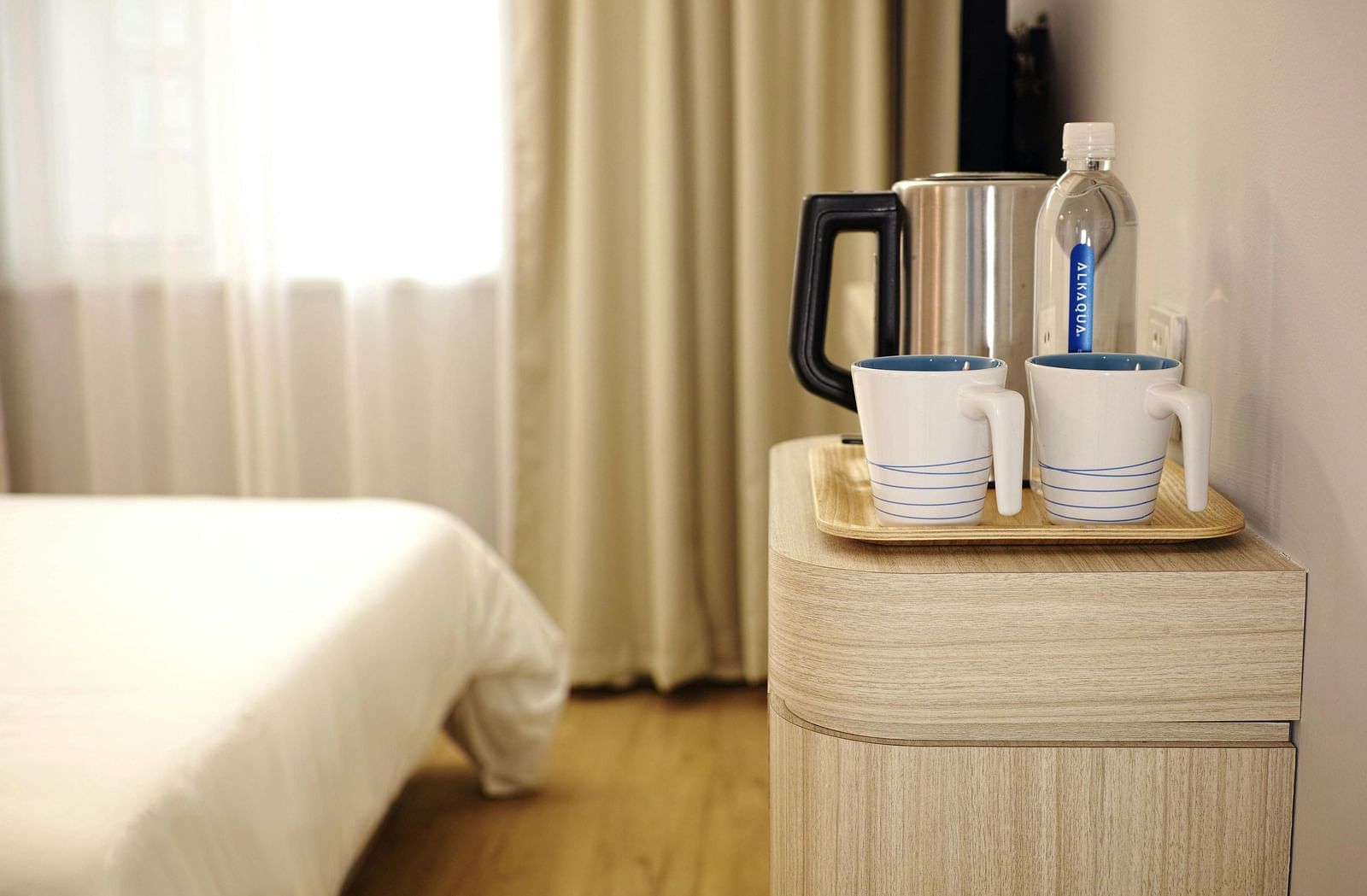 Two white ceramic mugs on brown wooden tray placed on a table next to a hotel bed