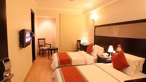 A spacious room at the Emblem Hotel New Friends Colony in New Delhi, featuring a large bed with a dark headboard, white linens, and pillows. A single bedside table with a lamp is visible next to the bed.