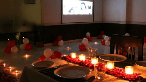 Romantic candlelit dinner setup at Polo Resort Cherrapunji with red rose petal runner, table for two and movie screen with balloons.