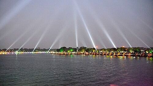 Night view of a lakeside promenade illuminated with vibrant lights and multiple white spotlights beaming into the sky, reflecting a festive atmosphere along the water's edge.