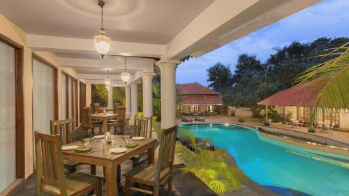 A poolside sit-out area at the resort featuring a wooden dining table set with plates and cutlery, overlooking the calm pool waters - Golden Tulip Goa Mandrem