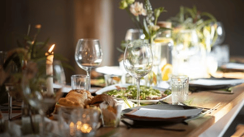 Candlelit dining table set with wine glasses, bread, greenery and small floral arrangements arranged for an intimate evening meal.