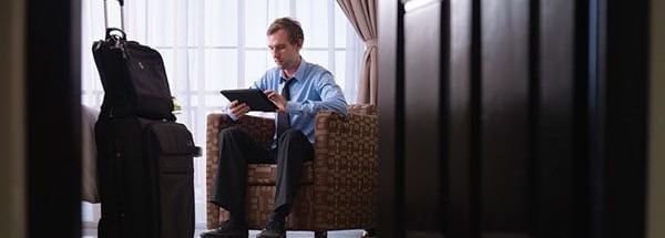 A man sitting on a sofa chair, with a suitcase and a small bag, beside a large window with the text “Find Hotel Near Mahabalipuram” showing.