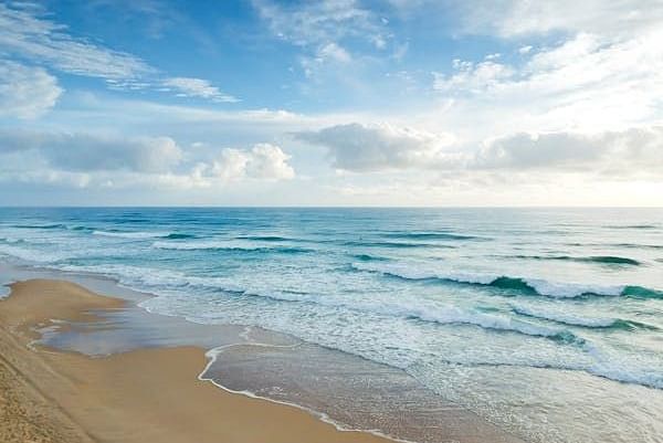 A wide coastal scene shows gentle waves rolling onto a sandy beach under a sky with scattered clouds.