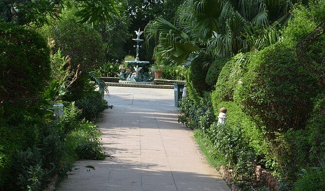A paved pathway leads through a lush green garden towards a fountain. 