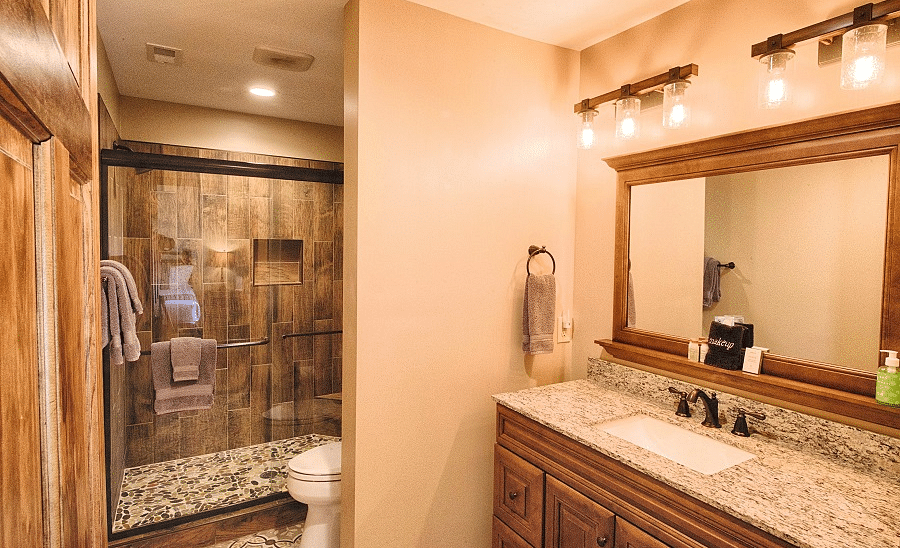 a bathroom with brown tiles featuring a washbasin with a wide wood framed mirror and a shower area - Historic Tapoco Lodge