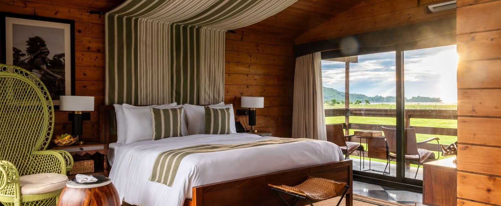 A bedroom with a rustic wooden interior, featuring a metal-framed bed with white bedding and a green striped runner. A large glass door opens to a balcony with a view of a grassy field and water. A woven stool and a jute rug add to the natural aesthetic. T