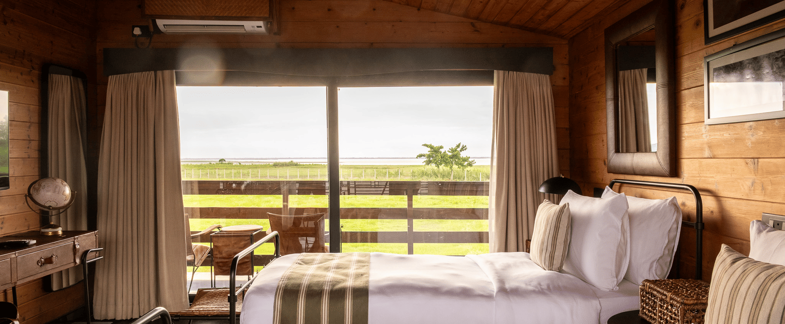 A bright and airy twin bedroom with two metal-framed beds, each with a white comforter and green striped runner. The room has wooden walls and a sloped ceiling. A large window provides a beautiful view of a grassy landscape and a body of water. This is a r