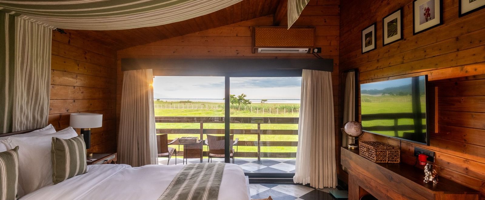 A wide shot of a bright bedroom with a bed featuring a striped canopy and a view of a lush green landscape through a large window. A wooden console holds a flat-screen TV and various decorative items. The floor is a striking black and white checkerboard ti