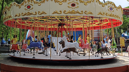 A colourful carousel with children riding ornate horse figures spins joyfully in a park under the shade of trees.