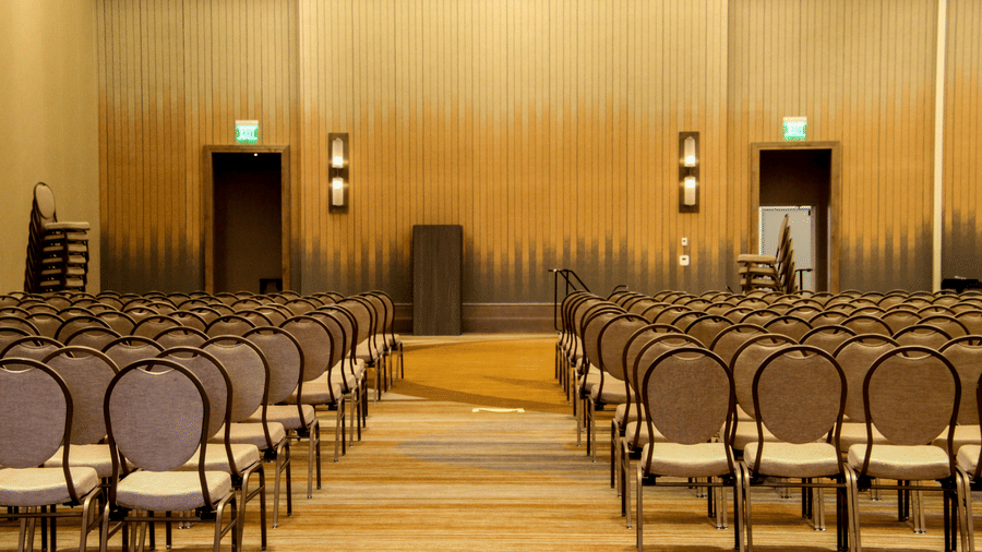 A large assembly room features rows of grey padded chairs facing a small wooden podium.