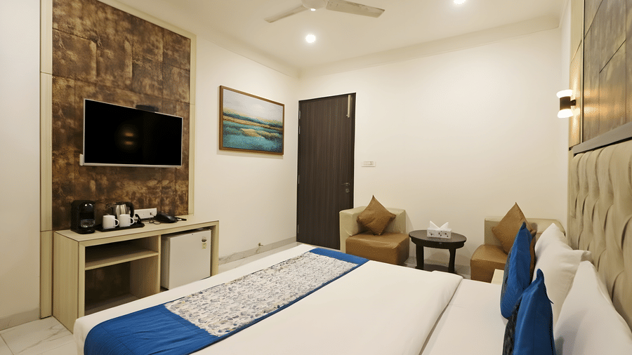 An interior view of a room at Hotel Apple Park Delhi showing a double bed, two tan armchairs with a small round table, and tea-making facilities on a desk.