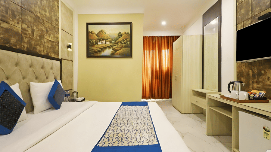A brightly lit superior room at Hotel Apple Park Delhi with a bed featuring blue cushions, a landscape painting on a lime green wall, and orange curtains.