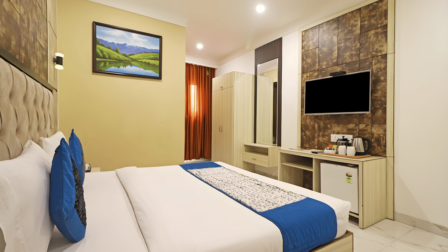 A perspective view of a king room at Hotel Apple Park Delhi showing a bed with blue accents, recessed ceiling lights, and a landscape picture above the headboard.