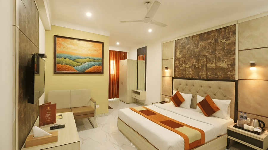A guest bedroom at Hotel Apple Park Delhi with a landscape painting above a seating area, a double bed with decorative cushions, and a ceiling fan.