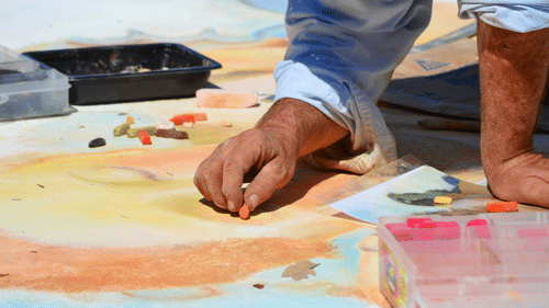 Two people’s hands working on a large painting with brushes and colors spread on a table.