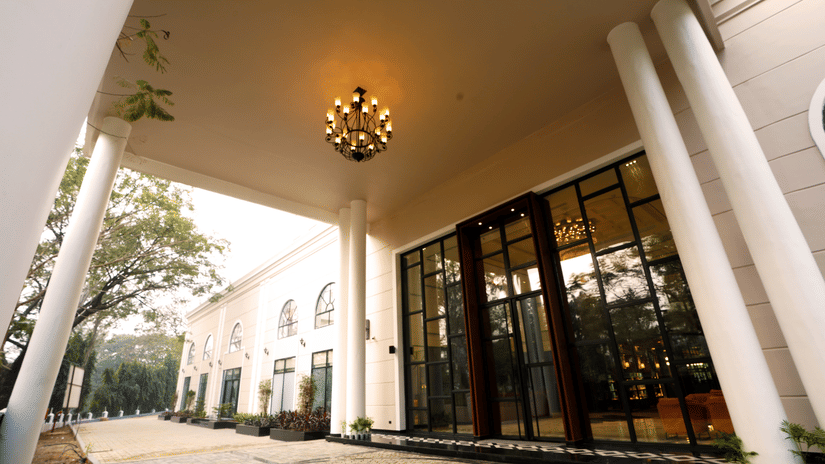 Hotel porte-cochere at Polo Hotel, Agartala with white columns, a tiered chandelier, glass entry doors and a circular mosaic medallion on the floor.