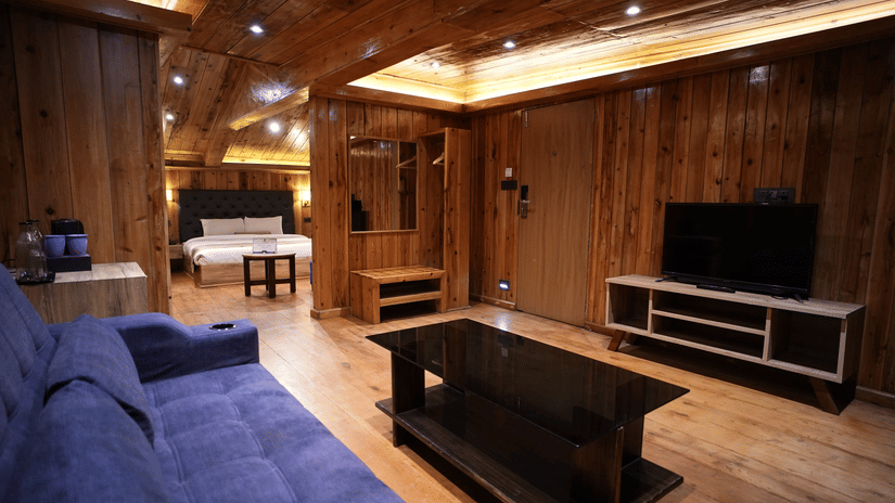 A spacious living area with panelled wood walls and ceiling, a seating sofa, a central coffee table, a television console, ceiling lights, and a double bed visible in the background at Mount Hotels.