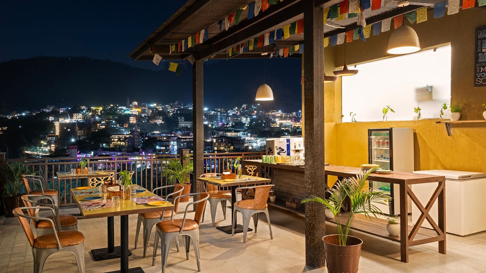 Panoramic view of city skyline from rooftop restaurant, with tables, chairs, and city lights at night at Bedzzz, Rishikesh 