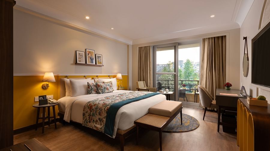 A spacious interior of room with a large bed, a sitting area with a sofa and a coffee table, and a desk. The room has large windows with curtains and a balcony overlooking a view.