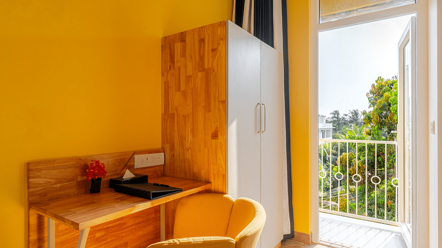 A cozy workspace in a room with yellow walls, featuring a wooden desk and chair set against a window with a view of foliage, creating a serene environment perfect for focus and creativity at Bedzzz Xclusiv Morjim, Goa