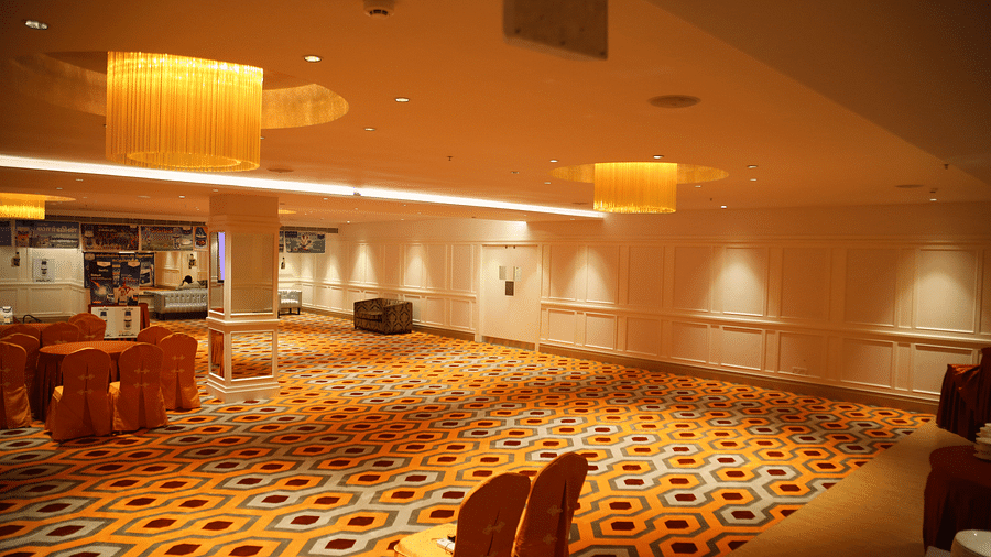 Large banquet room with geometric carpet, yellow pendant lighting and orange chair covers, with a stage in the background - TGI Grand Fortuna, Hosur