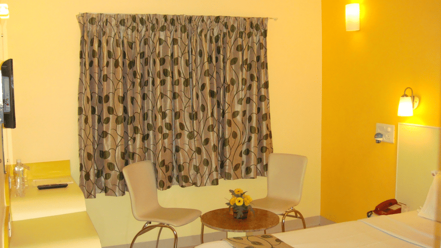 A clean hotel room with a single bed visible, a vibrant yellow corner wall, patterned window curtains, and 2 seating chairs - Hotel Pandian