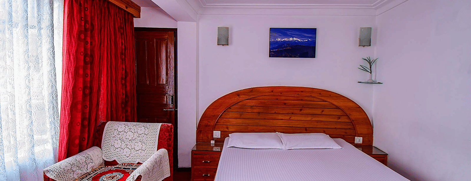 Close-up view of a double bed with a wooden headboard, red curtains, and 2 framed pictures on the wall at Hotel Dolphin Darjeeling.