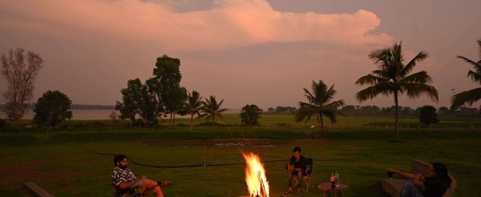 Campfire with chairs arranged around it on a grassy field, with a few people sitting and palm trees in the background.
