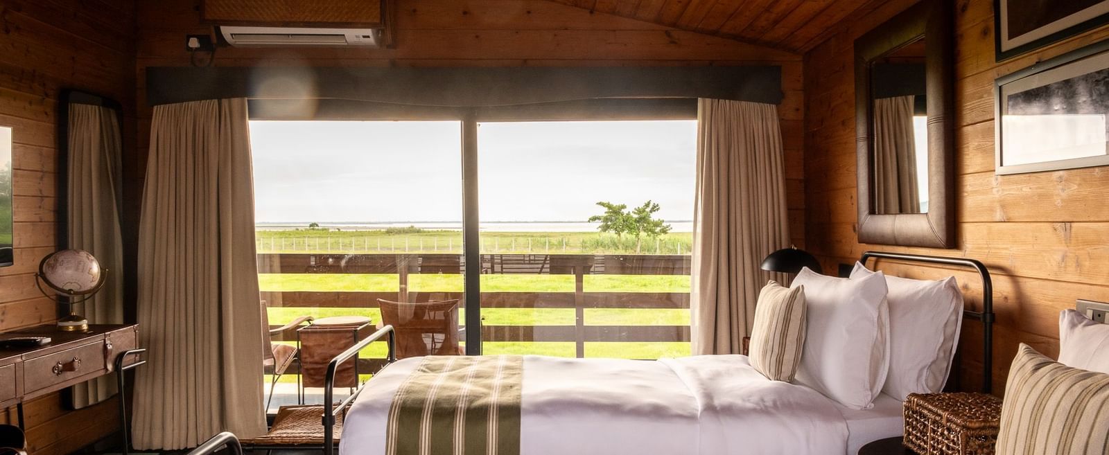 A bright and airy twin bedroom with two metal-framed beds, each with a white comforter and green striped runner. The room has wooden walls and a sloped ceiling. A large window provides a beautiful view of a grassy landscape and a body of water. This is a r