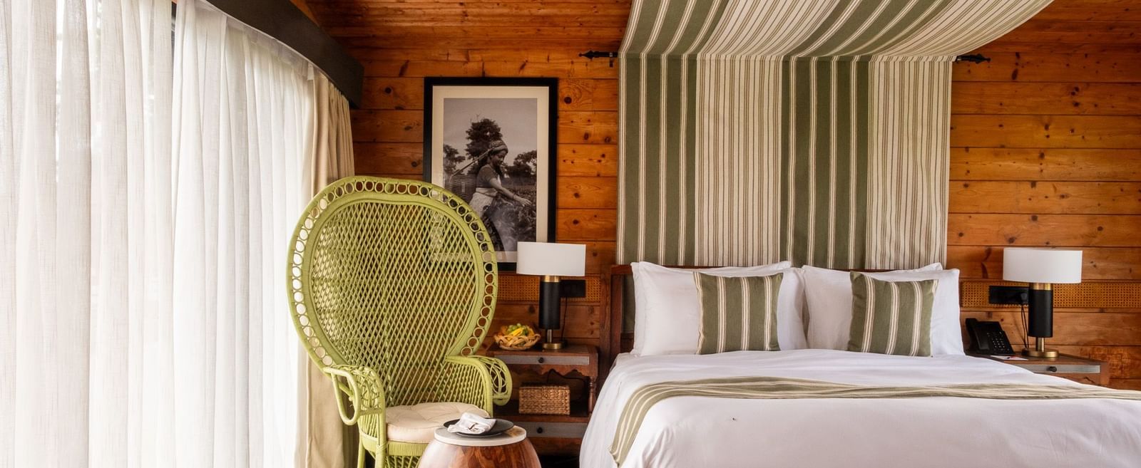 A rustic yet modern bedroom with a large bed, featuring a striped green and white canopy over the headboard. A bright green wicker peacock chair sits next to a large window with sheer curtains. The room has wooden paneled walls and ceiling, and a checkered