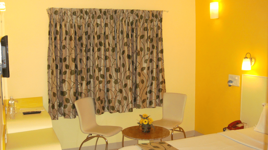 A clean hotel room with a single bed visible, a vibrant yellow corner wall, patterned window curtains, and 2 seating chairs - Hotel Pandian