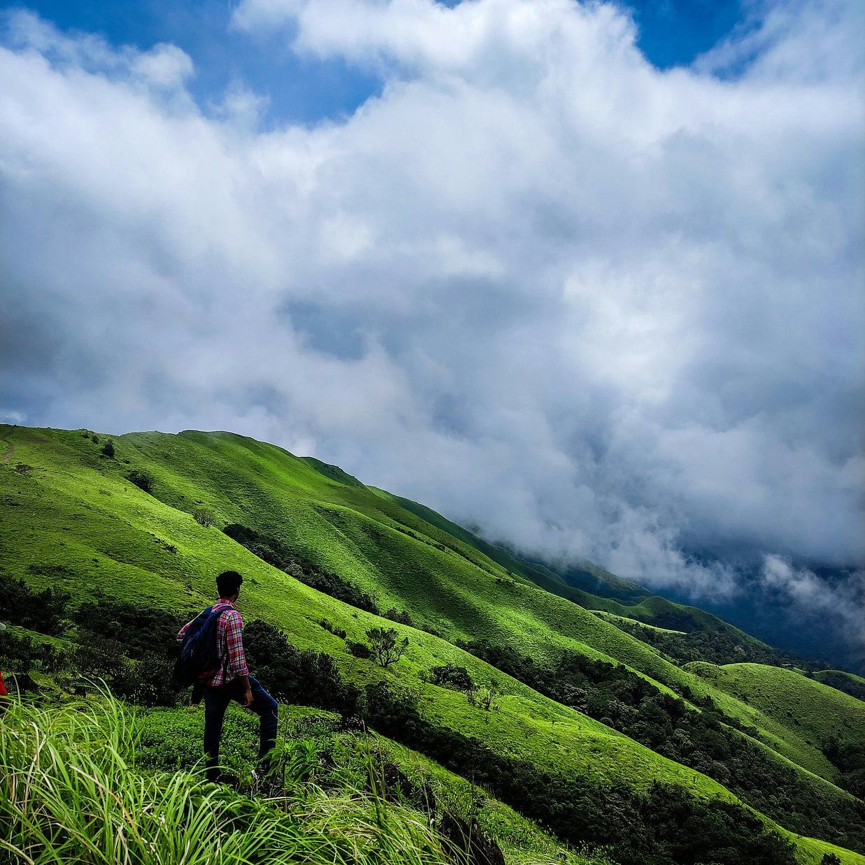 A lone trekker walking across a lush green hillside under a cloudy blue sky in a mountainous landscape.
