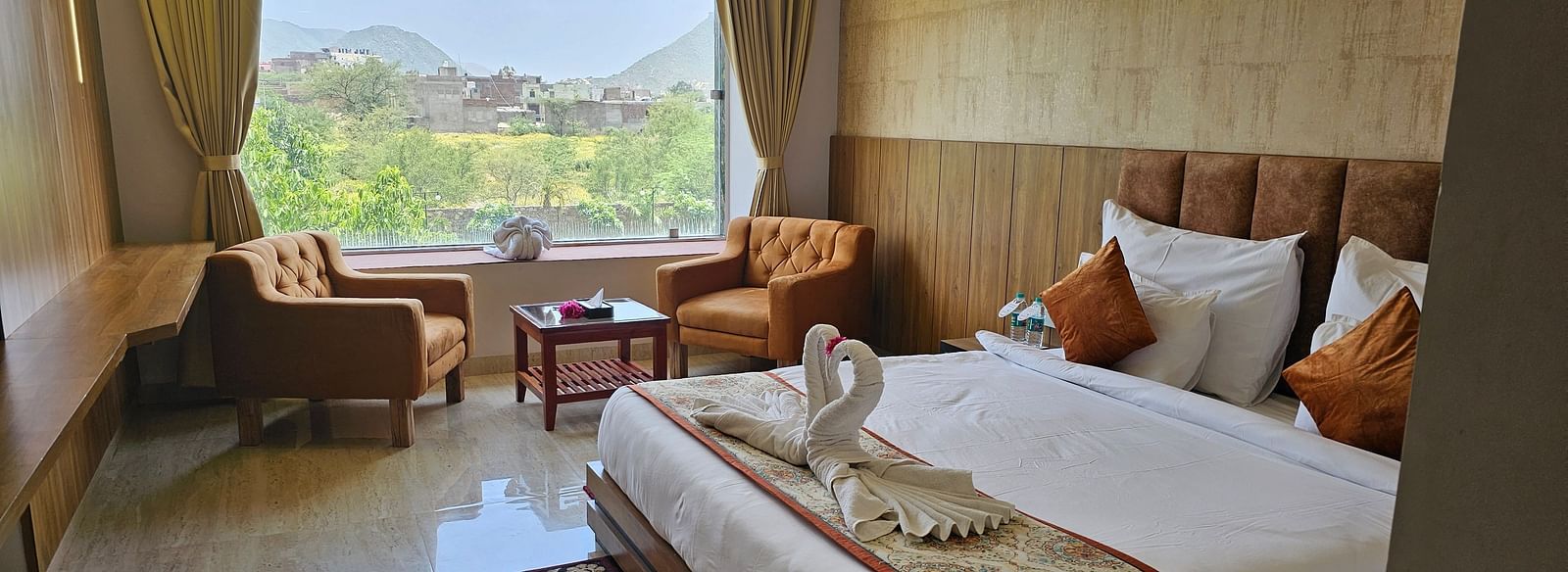 A bright and spacious resort room at Bamboo Saa Mulberry Resort, Pushkar, featuring a large window with a view of the outdoors including a made bed with a patterned runner, two armchairs, and a small table.