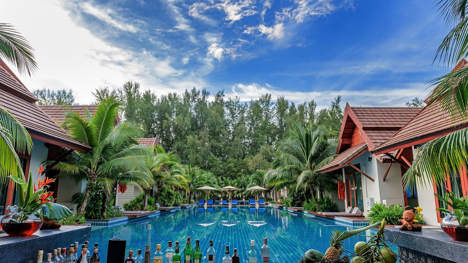 a swimming pool next to buildings on either side and trees covering the front with many drinks kept on the table