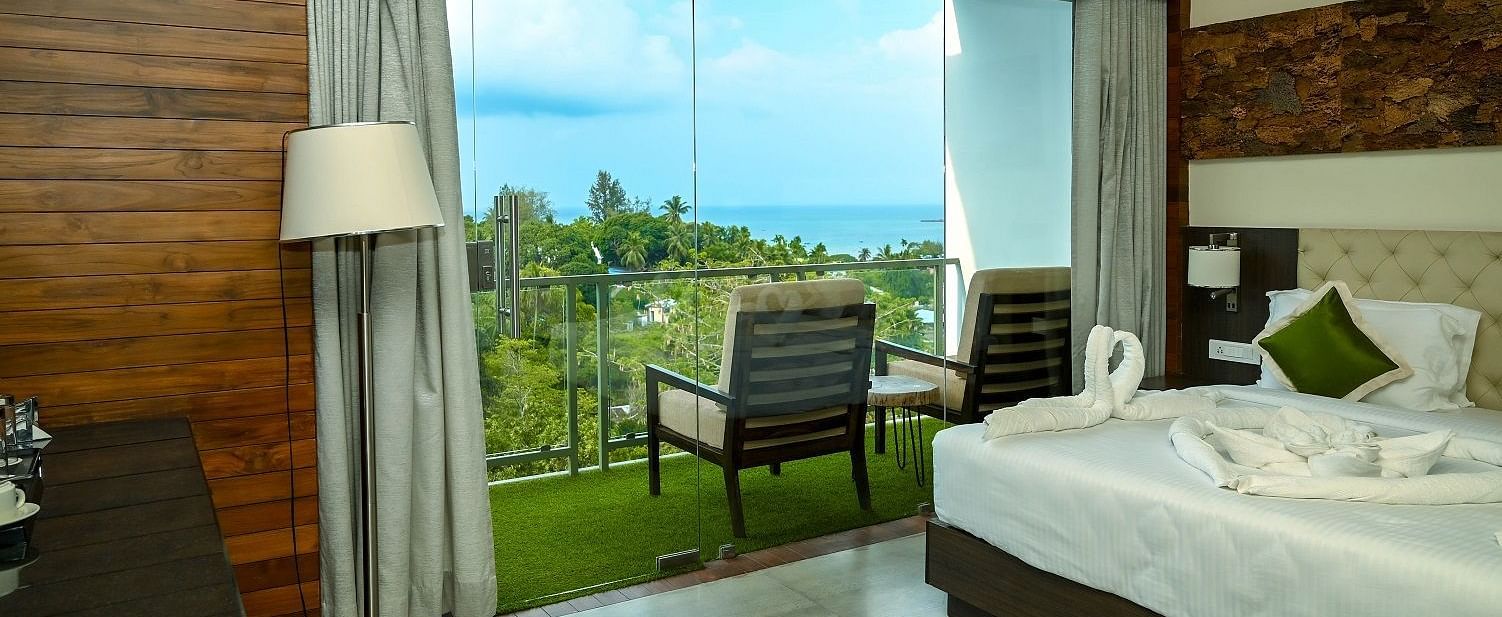 A hotel room interior featuring a large bed, a bedside lamp, and a full-length glass door leading to a balcony with an outdoor seating area, all against a backdrop of wooden wall panelling - Sea Hills Hotels, Sri Vijaya Puram (Port Blair)