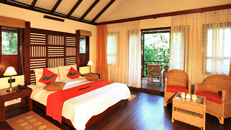 A bed placed in a room with 2 chairs and table, and a balcony that offers forest views at Vythiri Resort, Wayanad