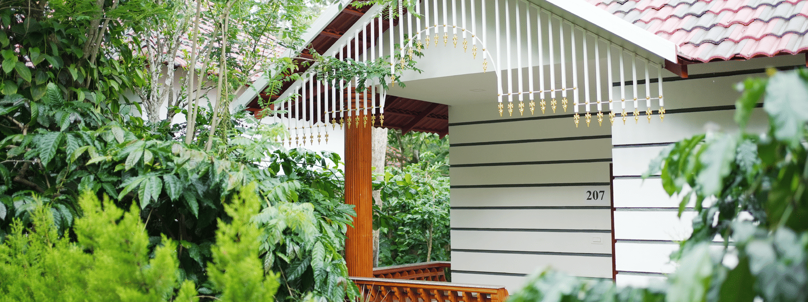 The exterior of a resort building with white horizontal siding, a red tiled roof, a covered porch, and dense surrounding greenery at Coorg Orange Blossom Resort and Spa.