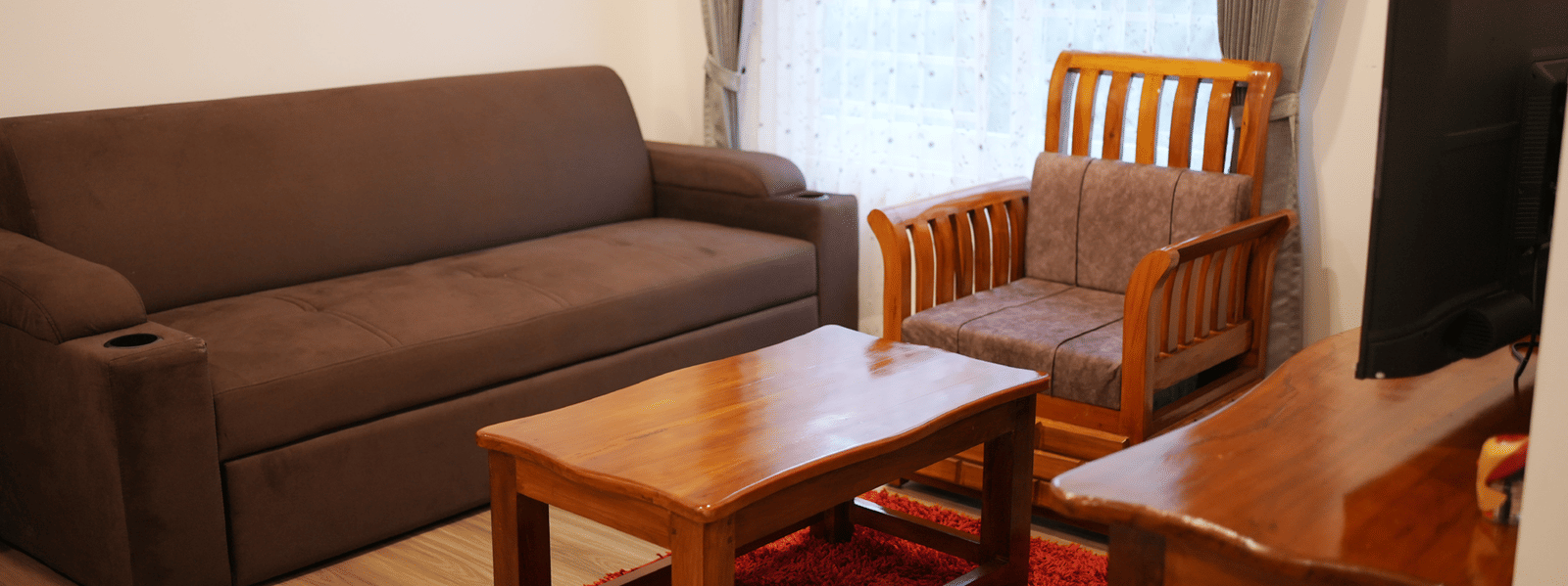 A cosy living room with a brown sofa, wooden chairs, coffee table, red rug, and television at Coorg Orange Blossom Resort and Spa.