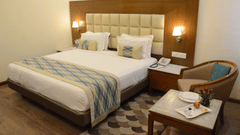 A well-appointed hotel room with a large bed, elegant wooden furnishings, and a cosy armchair in the corner - Clarks Avadh, Lucknow