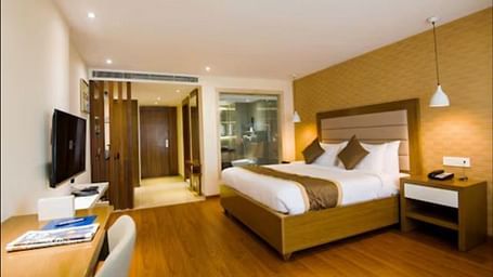 A deluxe hotel room with a large bed with wooden headboards, a bedside table, and a wall-mounted TV along with a wooden table below it at Yashshree The Imperial Inn, Siliguri.